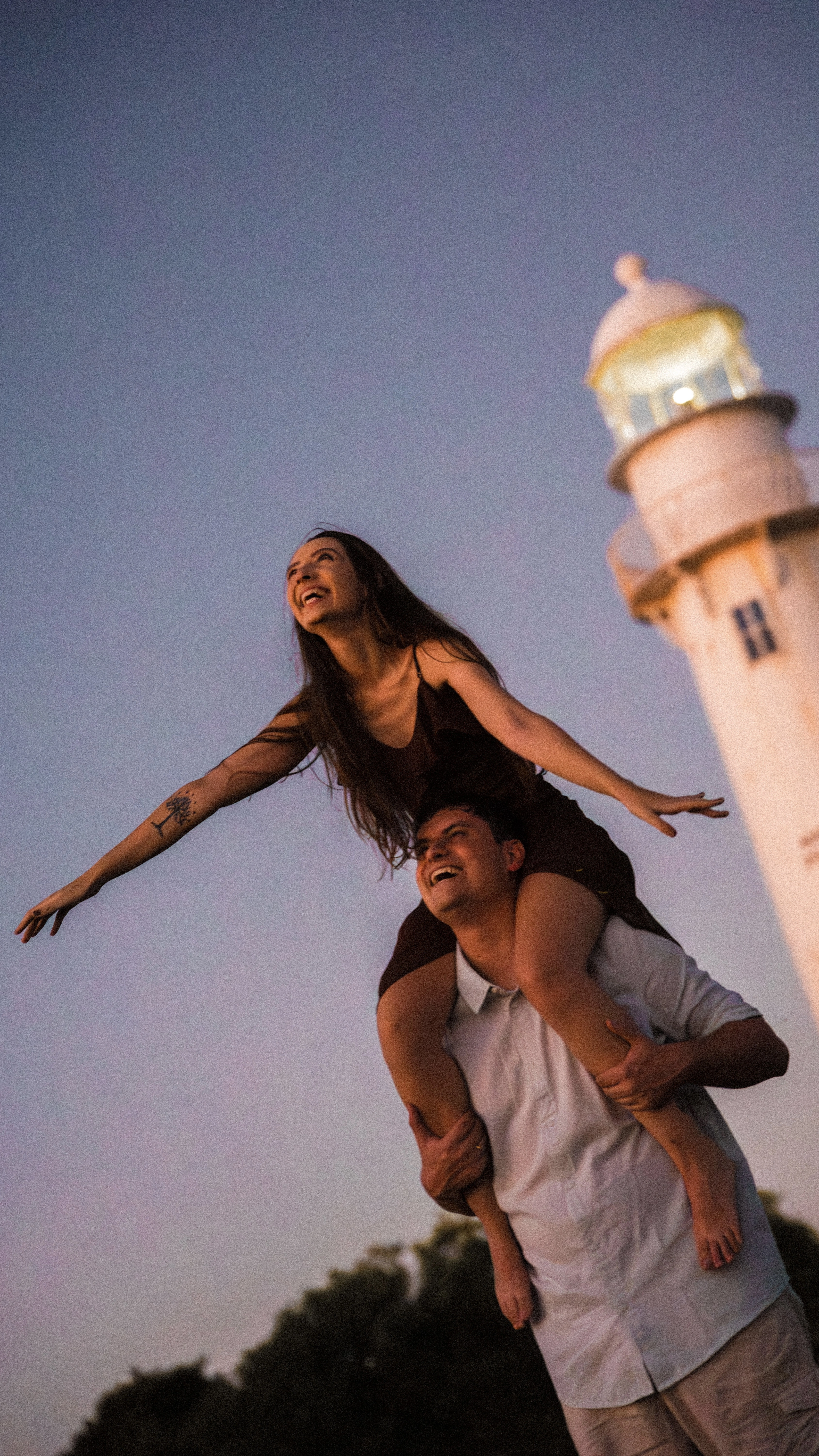 Os noivos estão juntos na praia durante o pôr do sol. A noiva sorri com as pernas apoiadas sobre os ombros do noivo. Ao fundo, aparece o farol da ilha.