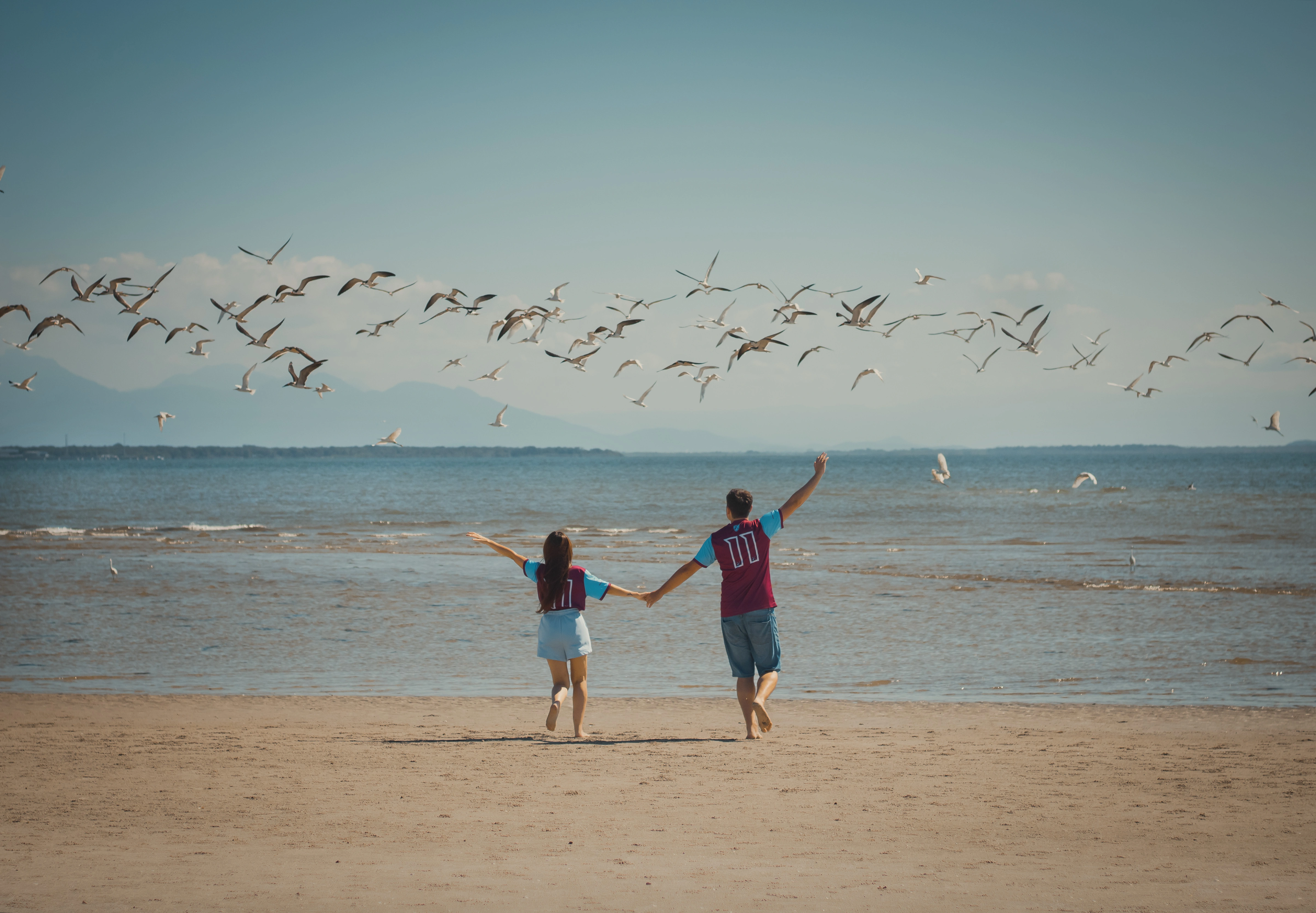 Os noivos estão na praia, descalços na areia, de mãos dadas e com os braços abertos, em pose de comemoração. Acima deles, vários pássaros voam pelo céu.