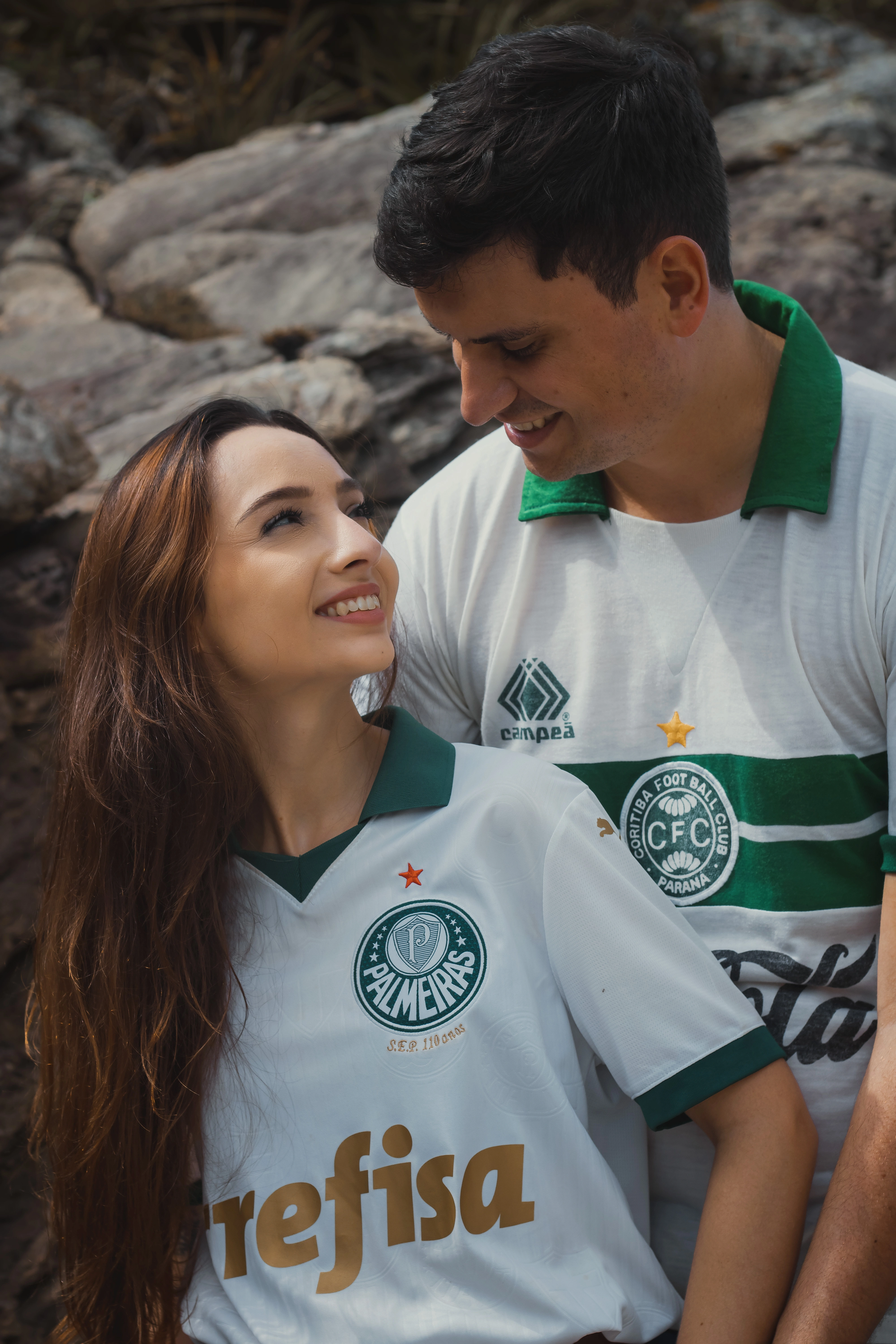 Os noivos se encaram sorrindo, com os rostos próximos. A noiva veste a camiseta mais recente do Palmeiras, enquanto o noivo usa uma camiseta retrô do Coritiba.