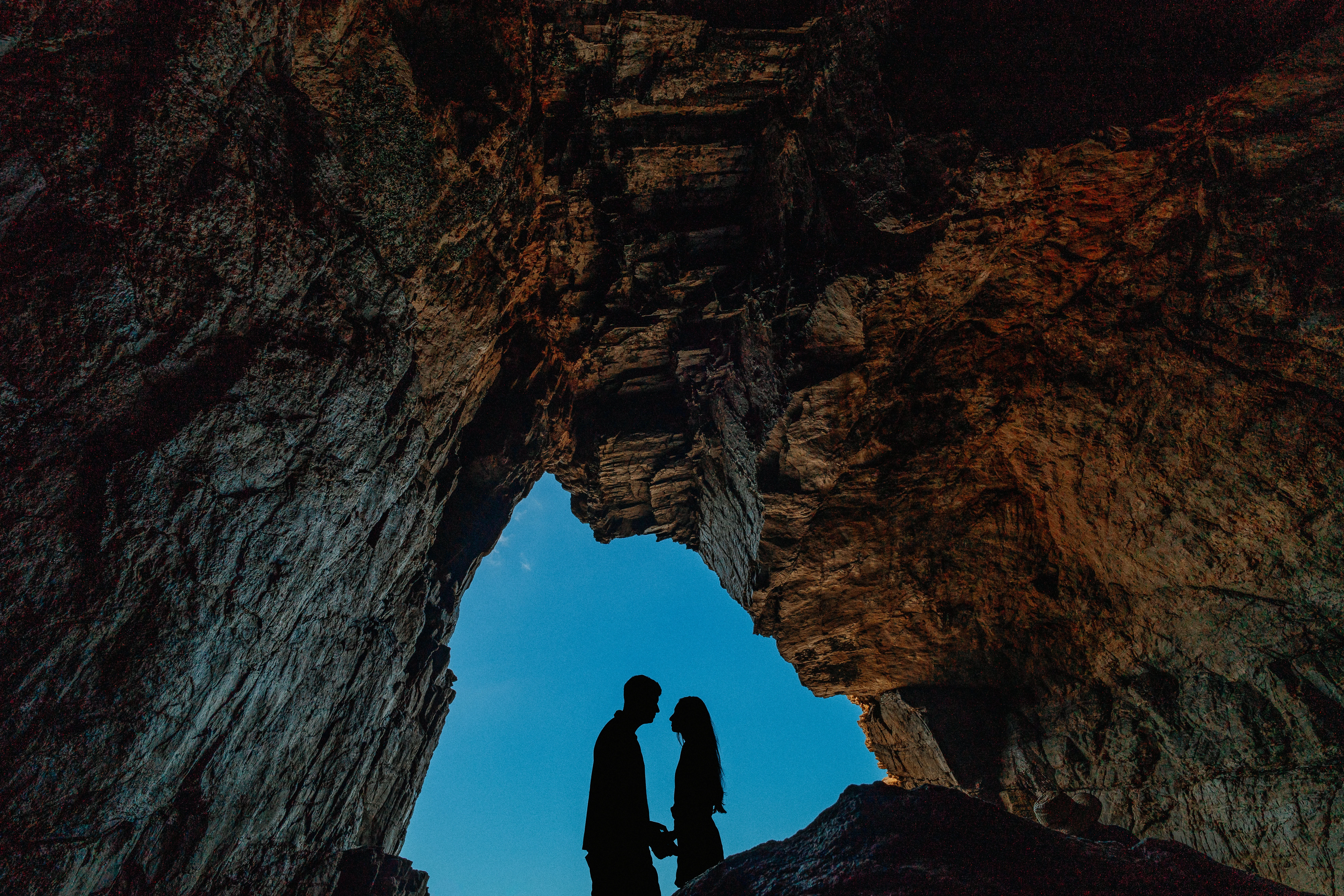 Os noivos estão na entrada de uma caverna, olhando um para o outro. A iluminação destaca apenas suas silhuetas.