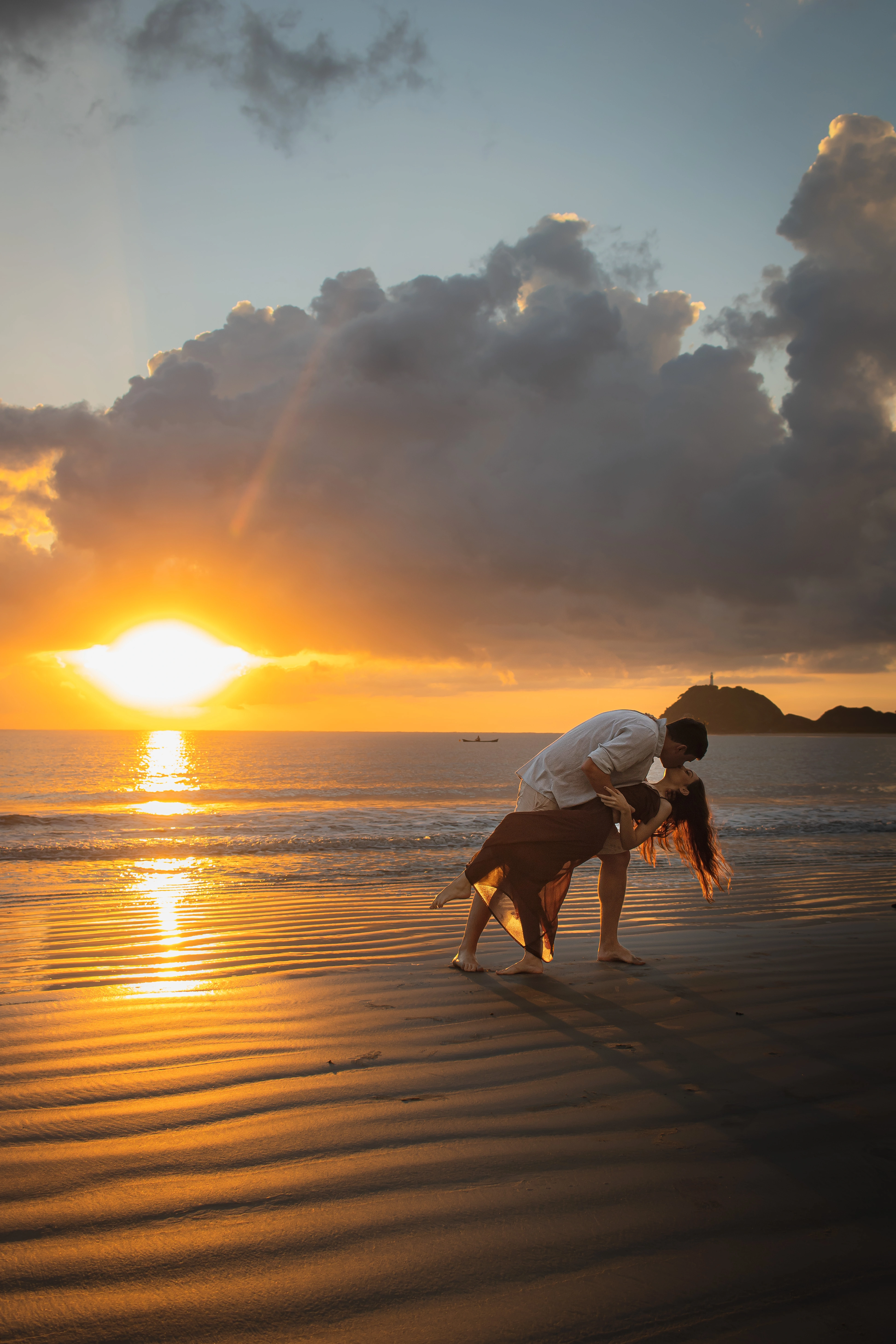 Os noivos se beijam na praia, próximos ao mar, com as ondas tocando seus pés. O noivo segura a noiva inclinada, sem deixá-la encostar na areia. Ao fundo, aparecem o pôr do sol e o farol da ilha.